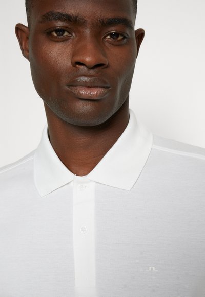 Camisa polo blanca con un acabado texturizado, que presenta un collar clásico y una tapeta de tres botones. Sutil acento del logo en el pecho.