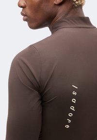 Gros plan d'une personne portant une chemise marron à manches longues avec « Isadore » imprimé verticalement sur l'épaule droite arrière.