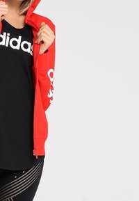Giacca rossa con zip e cappuccio, con logo bianco sul petto e manica; abbinata a una maglietta nera e leggings neri a motivo.
