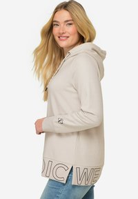 Haut à capuche beige avec une poche frontale, cordon de serrage, et ourlet fendu arborant l'imprimé "NORDIC WEAR" le long du bas. Manches longues et texture douce.