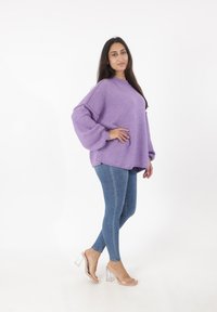 Lila übergroßer Pullover mit Puffärmeln, kombiniert mit blauen Skinny-Jeans und transparenten Blockabsatzsandalen. Glatte Textur.