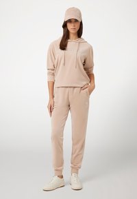 Felpa beige con logo "Guess" in strass, pantaloni jogger abbinati e un cappellino. L'outfit presenta una trama morbida e una vestibilità comoda. Sneakers bianche completano il look.