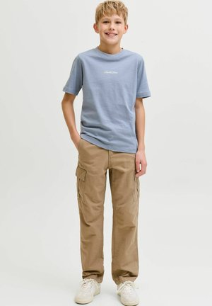 Naeratav poiss seisab käed taskutes, seljas helesinine T-särk, beežid püksid ja valged vabaajajalatsid, taustaks lihtne tasane pind.