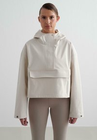 Veste courte beige clair avec capuche, présentant une poche avant, des fermetures à pression et des manches longues. Texture lisse avec un détail de marque subtil.