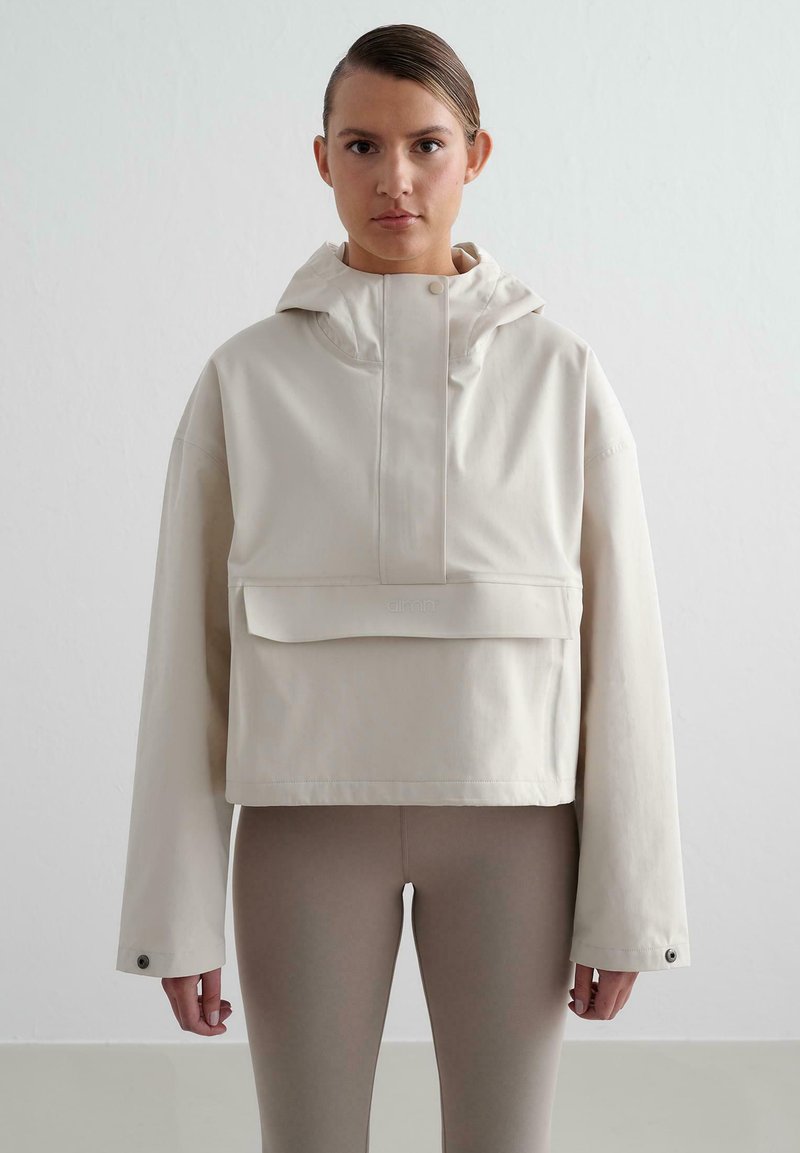 Veste courte beige clair avec capuche, présentant une poche avant, des fermetures à pression et des manches longues. Texture lisse avec un détail de marque subtil.