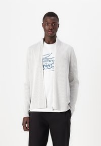 Svetlo šedý ryhovaný cardigan na zips s vystavaným golierom, nosený cez biele tričko s grafikou, v kombinácii s tmavými nohavicami.
