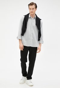 Lichtgrijze button-up shirt met opgerolde mouwen, gecombineerd met zwarte broeken en een zwarte trui over de schouders gedrapeerd. Witte sneakers maken de outfit af.