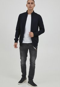 Giacca zip con collarino stand-up di colore blu navy, t-shirt bianca sotto, jeans grigi e sneakers nere. La giacca ha tasche con zip e un dettaglio con logo.