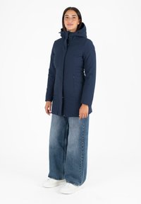 Parka isolante bleu marine avec capuche, dotée de boutons-pression et de poches latérales, portée sur un jean clair à coupe ample et des baskets blanches.
