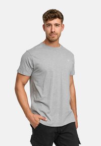 Graues T-Shirt mit kurzen Ärmeln und subtiler Textur, Rundhalsausschnitt und kleinem Logo-Detail. Getragen mit den Händen in den Taschen, kombiniert mit schwarzen Shorts.