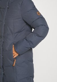 Mörkblå puffjacka med en texturerad yta, som har en logotyp patch på ärmen och en ficka med dragkedja och en brun dragflik.