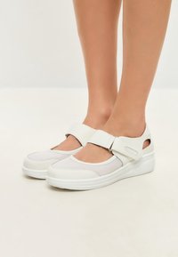 White shoes with a mesh upper featuring a rounded toe and adjustable strap. Thick sole provides elevation and comfort. Smooth texture.