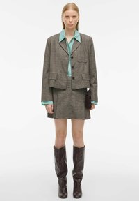 Bruine geruite blazer met een zak, gecombineerd met een turquoise blouse en minirok, en bruine kniehoge laarzen. Simpele bruine tas in de hand.