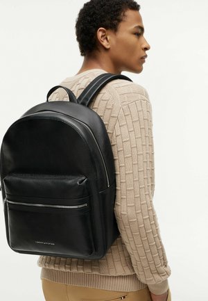 Persoon die een beige gebreide trui en beige broek draagt en een zwarte leren rugzak met een voorvak met rits bij zich heeft.