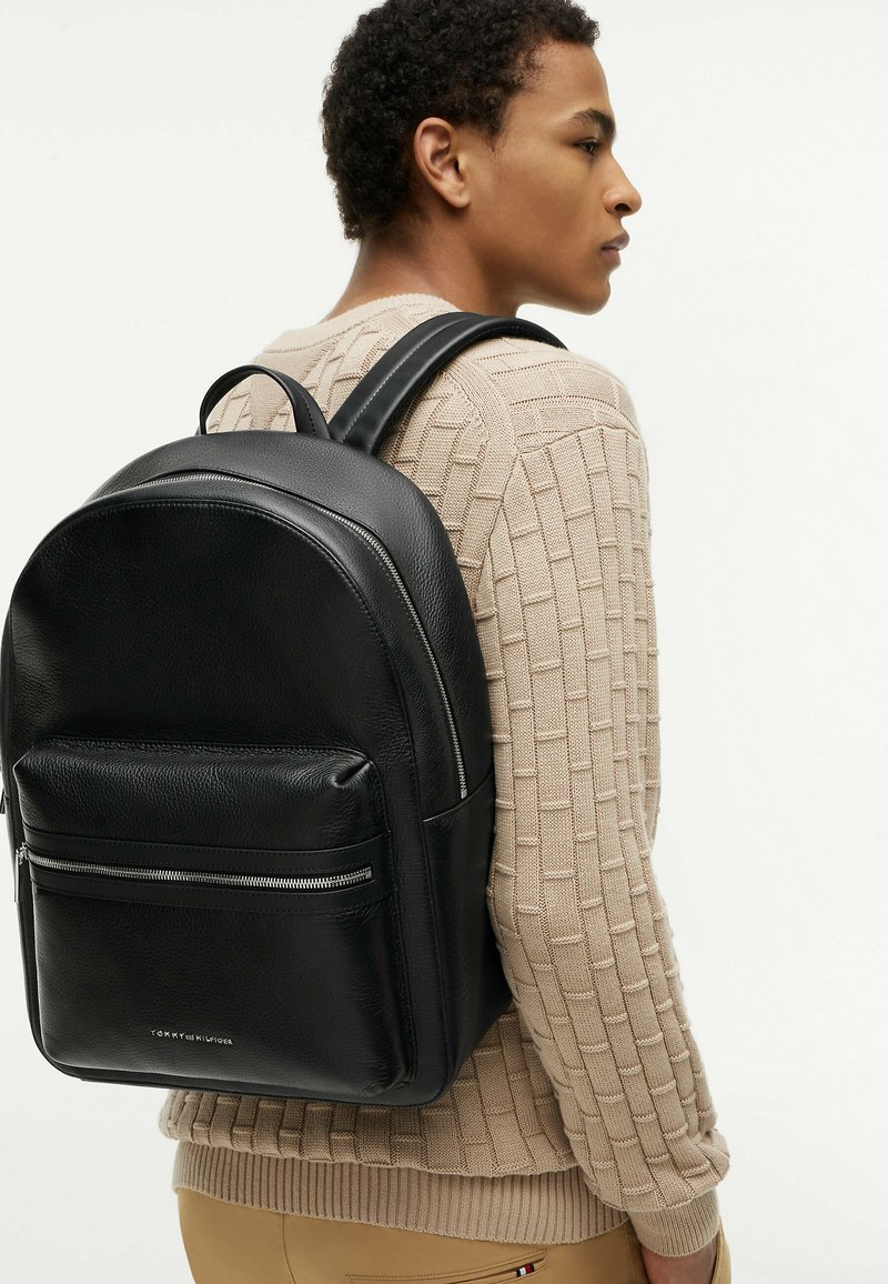 Personne portant un pull beige texturé et un pantalon beige, portant un sac à dos en cuir noir avec une poche zippée à l'avant.