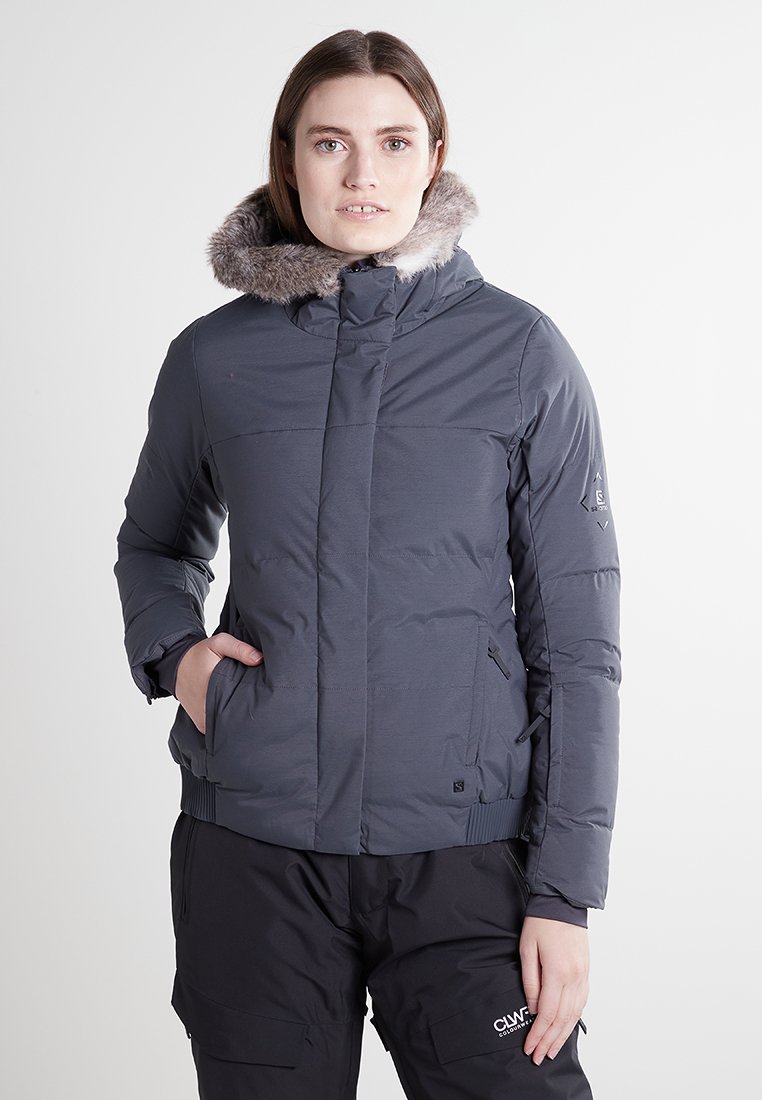 Chaqueta gris aislante con capucha forrada de pelo sintético, bolsillos con cremallera, puños acanalados y una textura suave. Presenta un diseño ajustado y un logo mínimo.