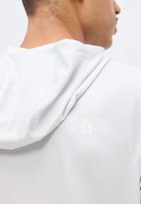 Homme portant un sweat à capuche blanc texturé avec le logo The North Face embossé sur la partie haute du dos, près de l'épaule.