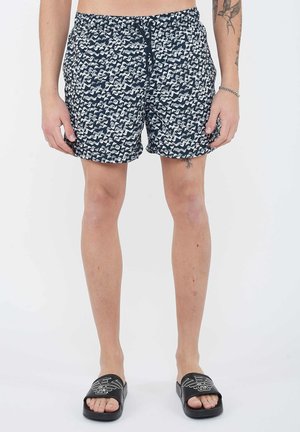 Uomo che indossa pantaloncini blu navy con fantasia e ciabatte nere, in piedi contro uno sfondo bianco, mostrando tatuaggi su braccia e gambe.