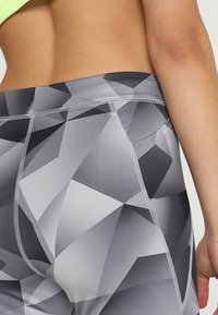 Lähikuva henkilöstä, jolla on päällä harmaat geometristen kuviiden leggingsit, joissa näkyy ompelutöitä ja sivutasku, jolloin osa heidän paljaasta selästään näkyy.
