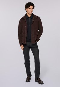 Brun mockaskinnjacka med huva, dragkedja framtill och fickor med lock, bärs över en svart skjorta med krage, ihop med mörkgrå jeans och bruna skor.