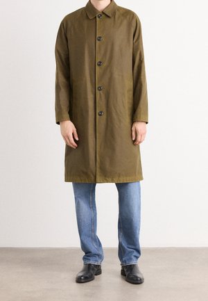 Homme portant un manteau vert olive longueur genou, un jean bleu et des bottines en cuir noir, debout devant un fond uni.