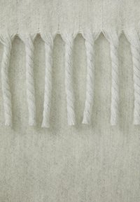 Tessuto grigio chiaro con una trama strutturata, caratterizzato da frange decorative realizzate con fili bianchi attorcigliati lungo il bordo inferiore.