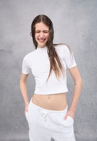 Weißes, kurzes T-Shirt mit kurzen Ärmeln und hohem Halsausschnitt, kombiniert mit weißen Jogginghosen mit Kordelzug, vor einem strukturierten grauen Hintergrund.