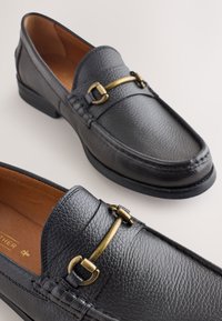 Mocassins en cuir noir avec une finition texturée, un accent en métal doré et un intérieur marron lisse. Talon bas empilé.