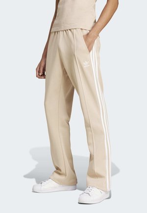 Persoon draagt beige Adidas trainingsbroek met witte strepen en logo, gecombineerd met witte sneakers, staand met één hand in de zak.