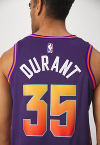 Homme portant un maillot de basket NBA violet avec "DURANT" et le numéro 35 dans le dos, debout devant un fond clair uni.
