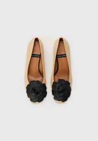 Ballerines beiges avec une texture lisse, arborant un grand détail en cuir noir en forme de fleur au niveau des orteils et une semelle intérieure brune avec marque.