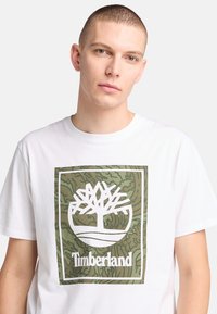 Camiseta blanca de algodón con un gran logo de árbol en verde y blanco con la palabra "Timberland" debajo. Cuello redondo simple y mangas cortas.