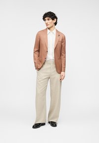 Ανοιχτό καφέ λινό σακάκι πάνω από λευκό πουκάμισο, συνδυασμένο με μπεζ παντελόνια φαρδιάς γραμμής και μαύρα παπούτσια· ομαλές υφές, μινιμαλιστικός σχεδιασμός, καθαρές γραμμές.