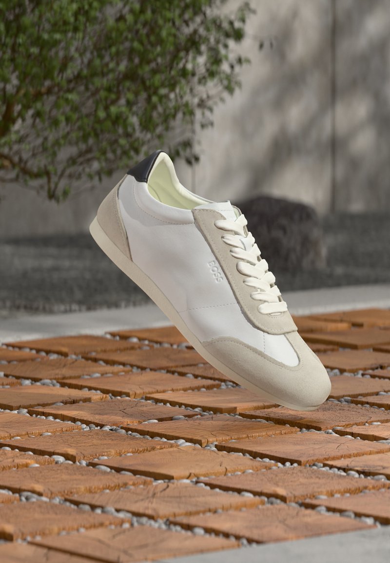Λευκό και μπεζ casual sneaker με μαύρη λεπτομέρεια στη φτέρνα, λευκά κορδόνια και λογότυπο "BOSS", να αιωρείται πάνω από ξύλινη πλακόστρωση σε εξωτερικό χώρο.