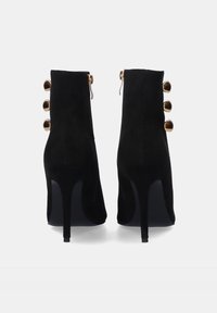 Botas de tobillo de ante negro con un alto tacón de aguja, que cuentan con un cierre de cremallera y seis botones decorativos en tono dorado en la parte trasera.
