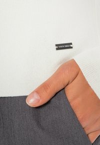 Biała dzianinowa bluzka o żebrowej fakturze, z czarną metalową metką z napisem "WITZIG" i szarym dołem z tkaniny. Częściowo widoczna ręka trzymająca materiał.