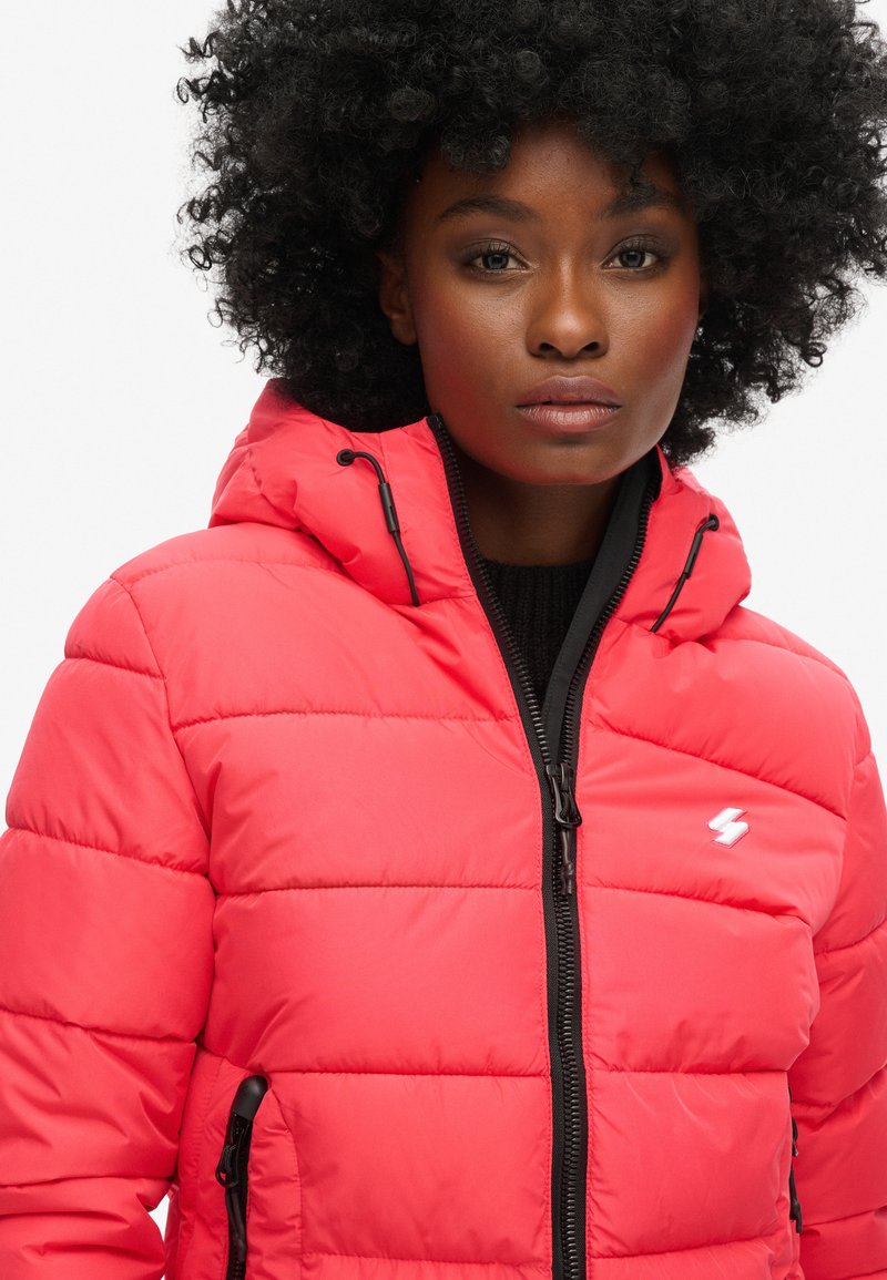 Czerwona kurtka puchowa z błyszczącym wykończeniem, poziomym pikowanym wzorem, zamkiem błyskawicznym z przodu oraz czarnymi akcentami na mankietach i dolnej krawędzi.