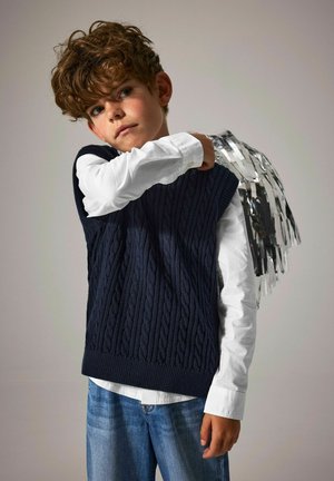 Jongen met krullend haar, gekleed in een witte shirt en een marineblauwe gebreide vest, met een zilverkleurige metallic franjeversiering over zijn schouder.