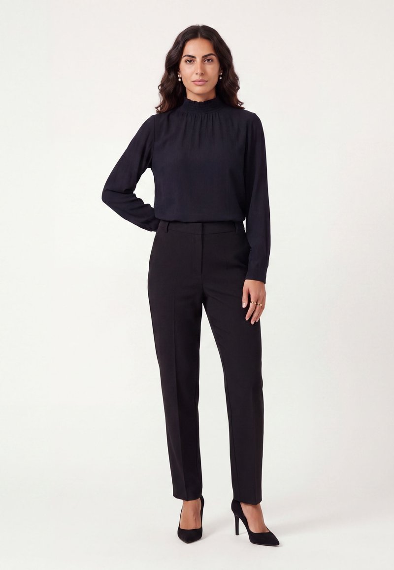 Woman standing against plain background wearing a black long-sleeve blouse, black trousers, and black high heels.