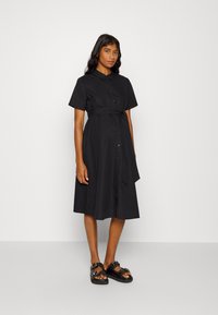 Vestido preto de botão, com mangas curtas, cintura cintada e saia até ao joelho. Usado com sandálias pretas com tachas. Textura de tecido suave.