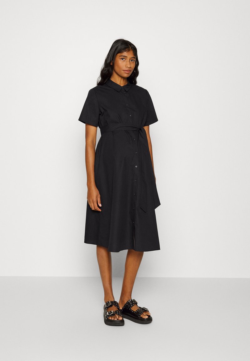 Vestido preto de botão, com mangas curtas, cintura cintada e saia até ao joelho. Usado com sandálias pretas com tachas. Textura de tecido suave.