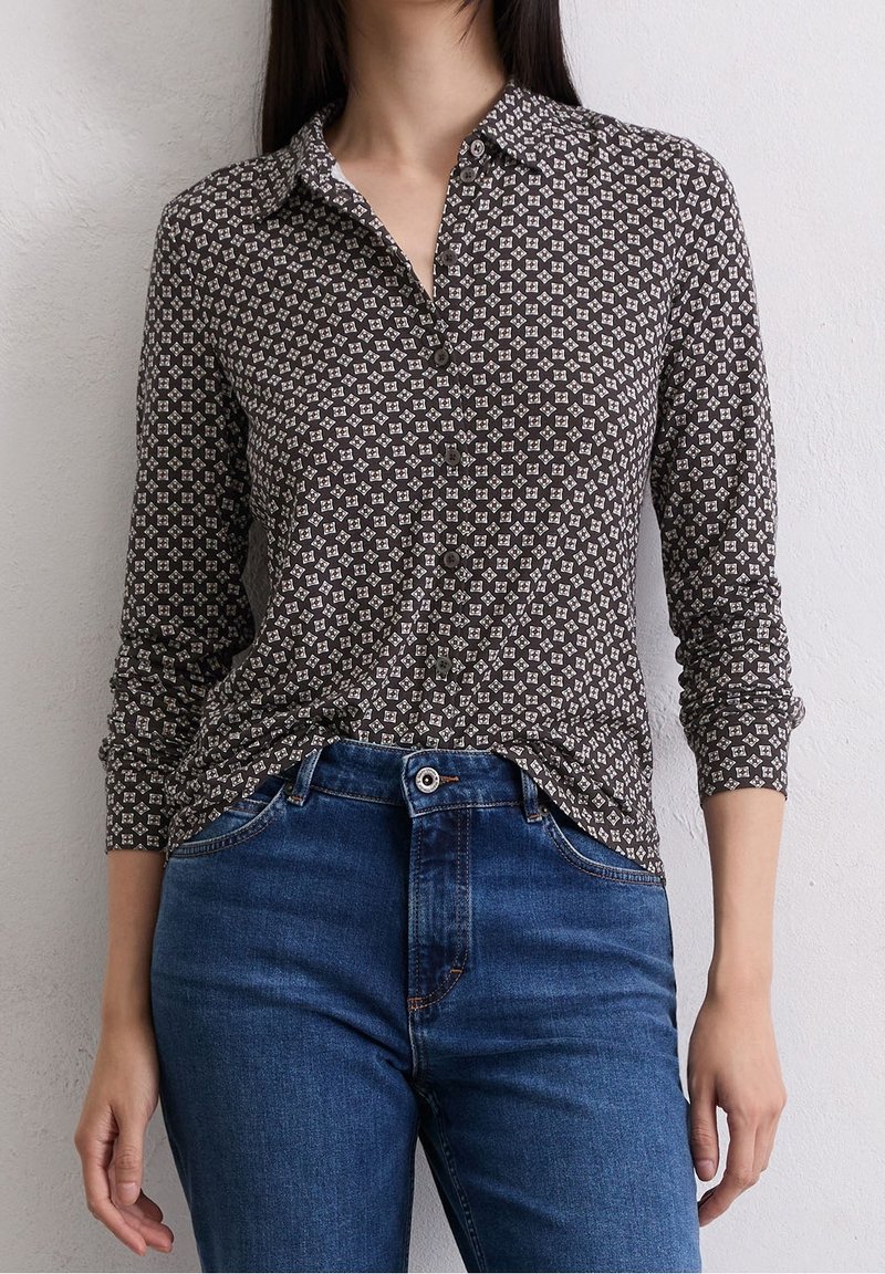 Femme portant une chemise marron à motif à manches longues boutonnée, rentrée dans un jean bleu taille haute, debout contre un mur blanc uni.