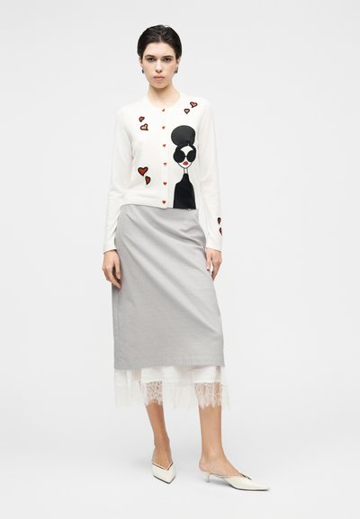 Fehér kardigán fekete és piros szív- és arc mintával, szürke szoknyával, amely csipke díszítéssel rendelkezik. A modell magassarkúban.