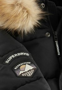Melna pildīta jaka ar noņemamu mākslīgās kažokādas kapuci, ar Superdry logotipu un "Expedition Everest" uzšuvi uz piedurknes.