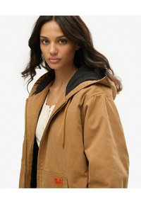Brązowa kurtka z kapturem z zamkiem błyskawicznym, czarnym podszyciem i naszywką na przedniej kieszeni. Wykonana z trwałego materiału o miękkiej teksturze.