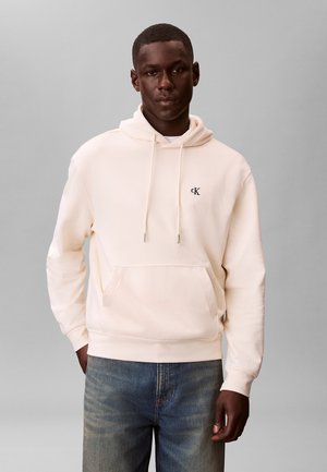 Hombre con una sudadera con capucha color crema con bolsillo frontal y el logo de CK en el pecho, combinado con unos vaqueros azules desgastados, de pie frente a un fondo gris liso.