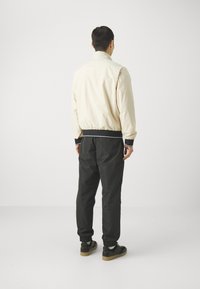 Bomber jacket beige chiaro con colletto e polsini elasticizzati, abbinata a pantaloni neri. Texture liscia, design minimalista, accenti bianchi a contrasto.