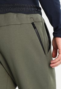 Gröna sweatpants med svart elastiskt midjeband, med en zipficka på sidan, tillverkade av ett mjukt tyg med en slät yta.