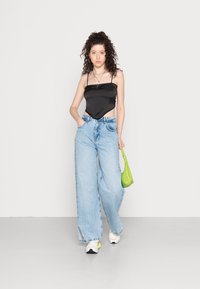 Camisola de cetim preta com alças finas, combinada com jeans claros de perna larga. Segurando uma pequena bolsa verde, ténis brancos completam o look.