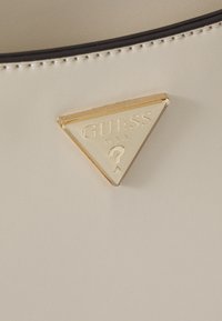 Materiale sintetico di colore crema con una finitura testurizzata, dotato di un logo triangolare dorato in rilievo con la scritta "GUESS U.S.A." e un punto interrogativo.